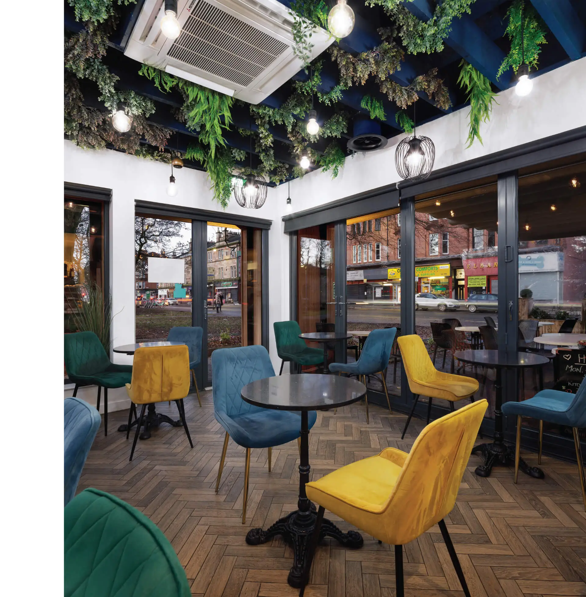 An image of the interior of a modern commercial café with colourful chair in yellow, blue and green, round tall tables, hardwood herringbone floor, and dark blue ceiling rafters between which plants are seen hanging. The space is bright, the walls are white and the large floor-to-ceiling windows reveal a lot of the streets outside like a painting. This project was designed by NVDC architects specialists in commercial and retail design.