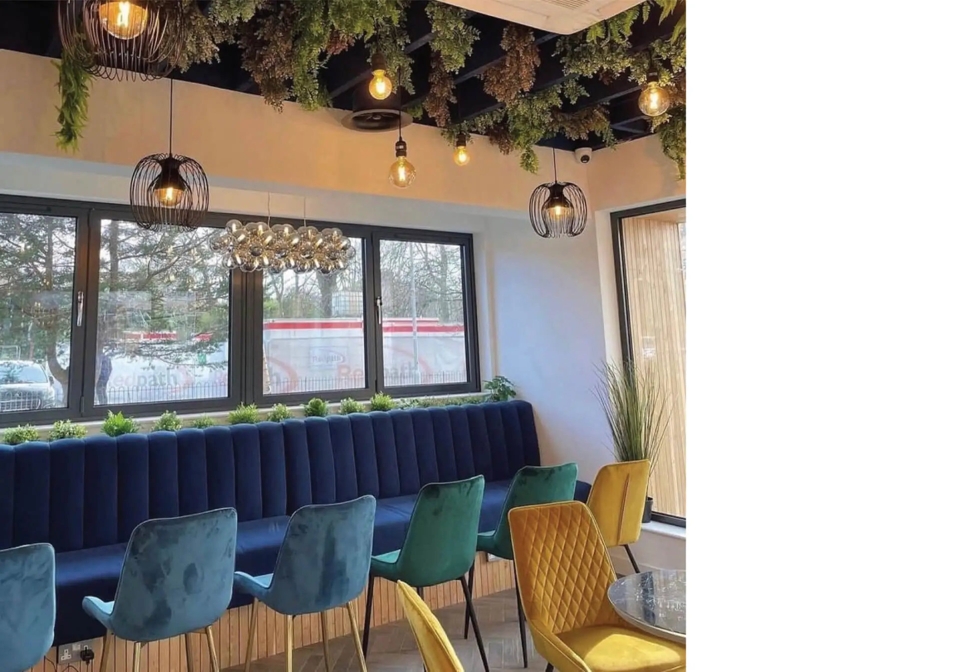 An image of the interior of a modern commercial café with colourful chair in yellow, blue and green, blue velvet booth seating, round tall tables, hardwood herringbone floor, and dark blue ceiling rafters between which plants are seen hanging. This project was designed by NVDC architects specialists in commercial and retail design.