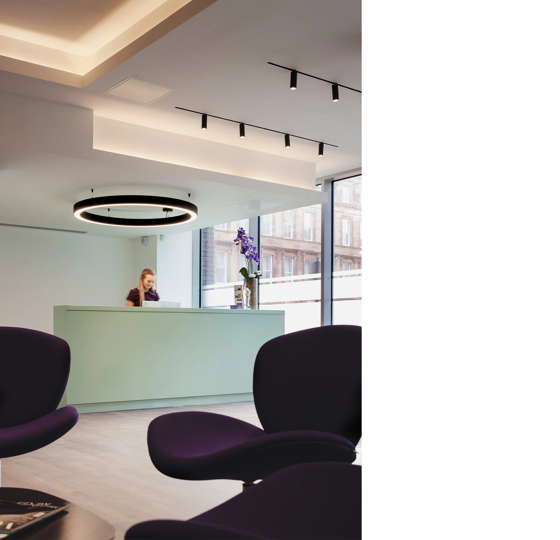 Interior of hospital reception with white ceiling with LED lights, light green reception desk, floor-to-ceiling windows providing natural light and dark purple seating in the foreground designed by award-winning healthcare architects.