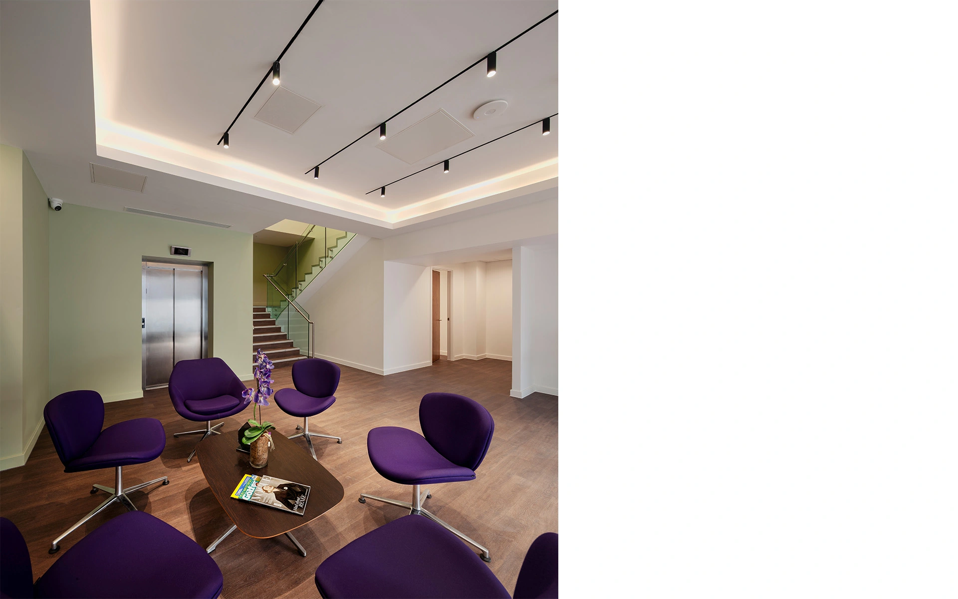 Interior of hospital reception with white ceiling with LED lights, dark purple seating and stairs with glass railing and timber floor.