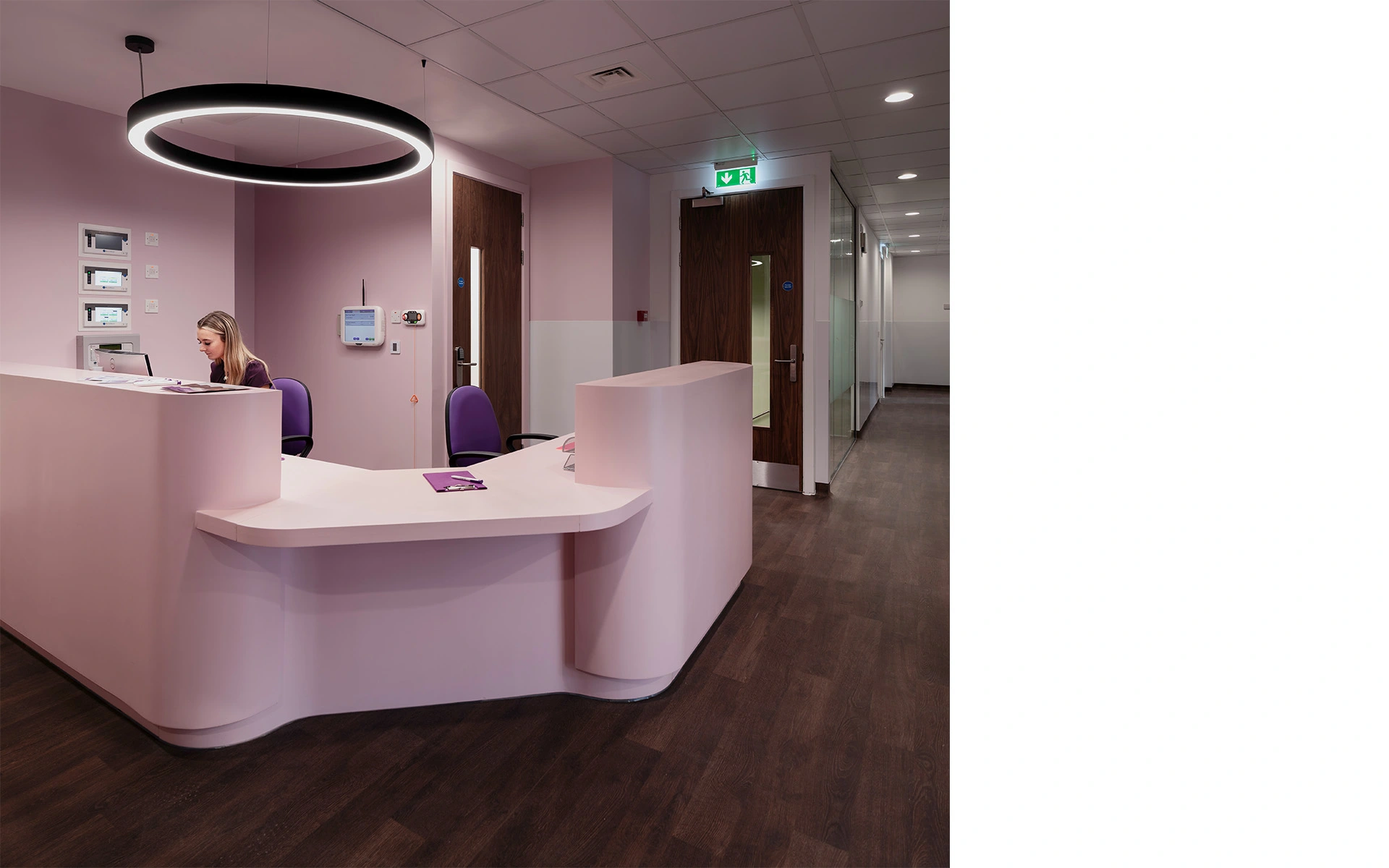 Reception desk in light pink on the second floor of specialised hospital premises in Glasgow, with dark timber flooring, contemporary circle lights and glass partition walls allowing natural light into the stairwell.