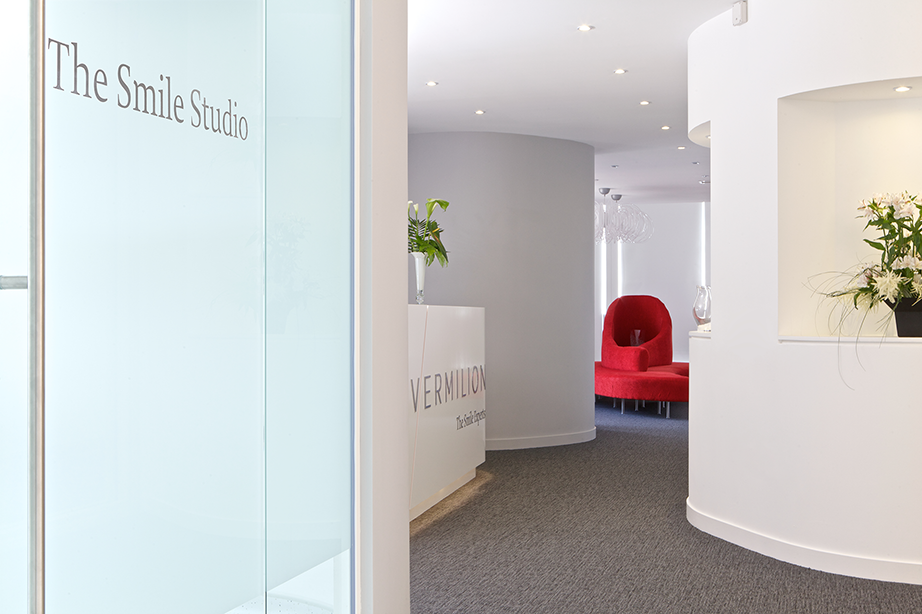 Stylish and modern dental practice interior designed by a specialist healthcare architect, featuring a curved reception desk and red accent seating. The dental design includes a bright, welcoming entrance, ideal for healthcare and medical practices aiming to provide a professional yet comfortable patient experience. The matted partition glass door on the left reads "The Smile Studio" and a statement red chair is visible in the middle of the waiting area, where patient comfort is priorities with large windows for natural light.