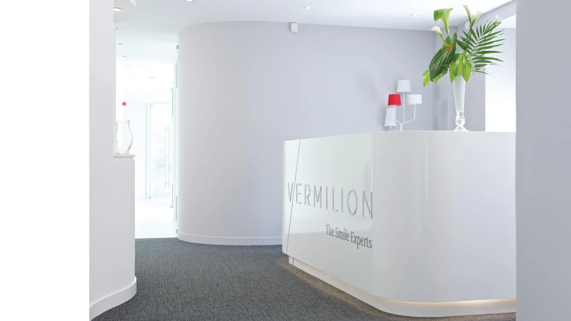 Reception area of a modern dental practice designed by healthcare specialist architects NVDC, featuring sleek white surfaces, minimalist décor, and bright lighting. The design showcases a professional and welcoming space with specialist dental practice design elements, including a curved reception desk and plant arrangements. Right behind the reception desk the entrance to a bright hallway is visible leading to the dental surgeries.