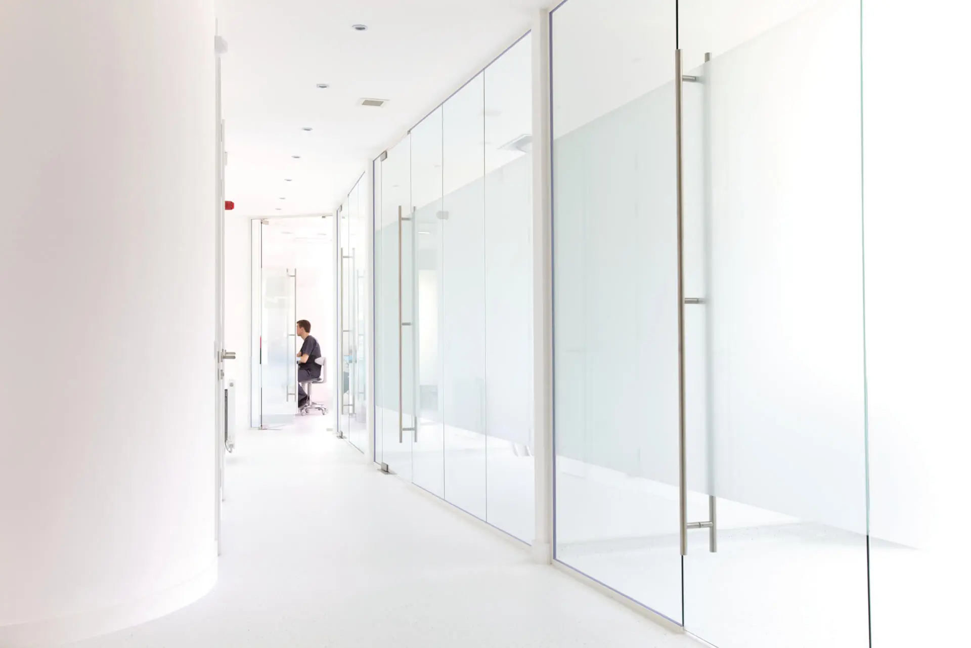 Bright and modern corridor of a dental practice with floor-to-ceiling glass partitions, designed by healthcare architects. The transparent medical facility design emphasizes cleanliness and openness, essential for healthcare environments. This dental design layout highlights efficient use of space, offering privacy while maintaining a welcoming atmosphere. This project was designed by NVDC healthcare and dental practice design specialists Glasgow.