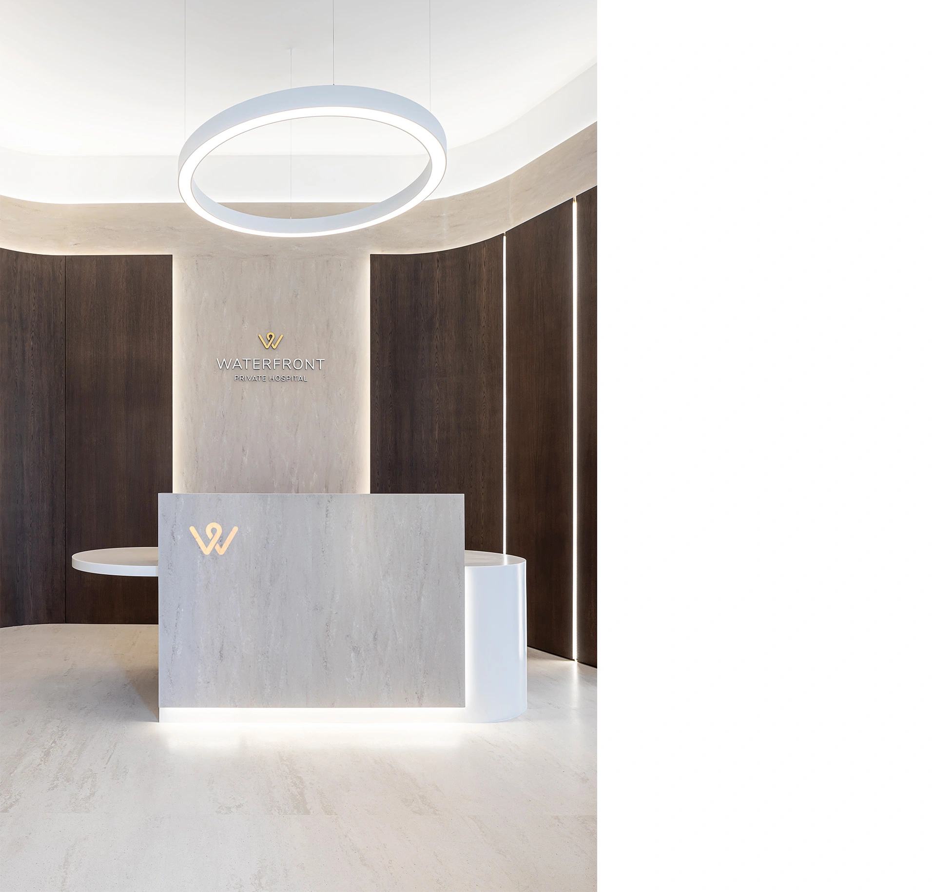 A photo of the front view of a modern reception waiting space with polished white stone floors and walls, white ceiling with curved long light features guiding the way to the stairs and LEDs between the dark timber panels layered on top of the stone on the wall. The reception desk is also designed in the same blonde stone material and the elegant signage reading "waterfront Private Hospital" stands out on the smooth and polished surfaces of the walls and reception desk. Below the reception desk there are LEDs creating an effect to make it look like the desk is floating. This bespoke modern hospital waiting space was designed by NVDC healthcare specialist medical architects who have almost two decades of experience with clinic and healthcare architecture premises design.