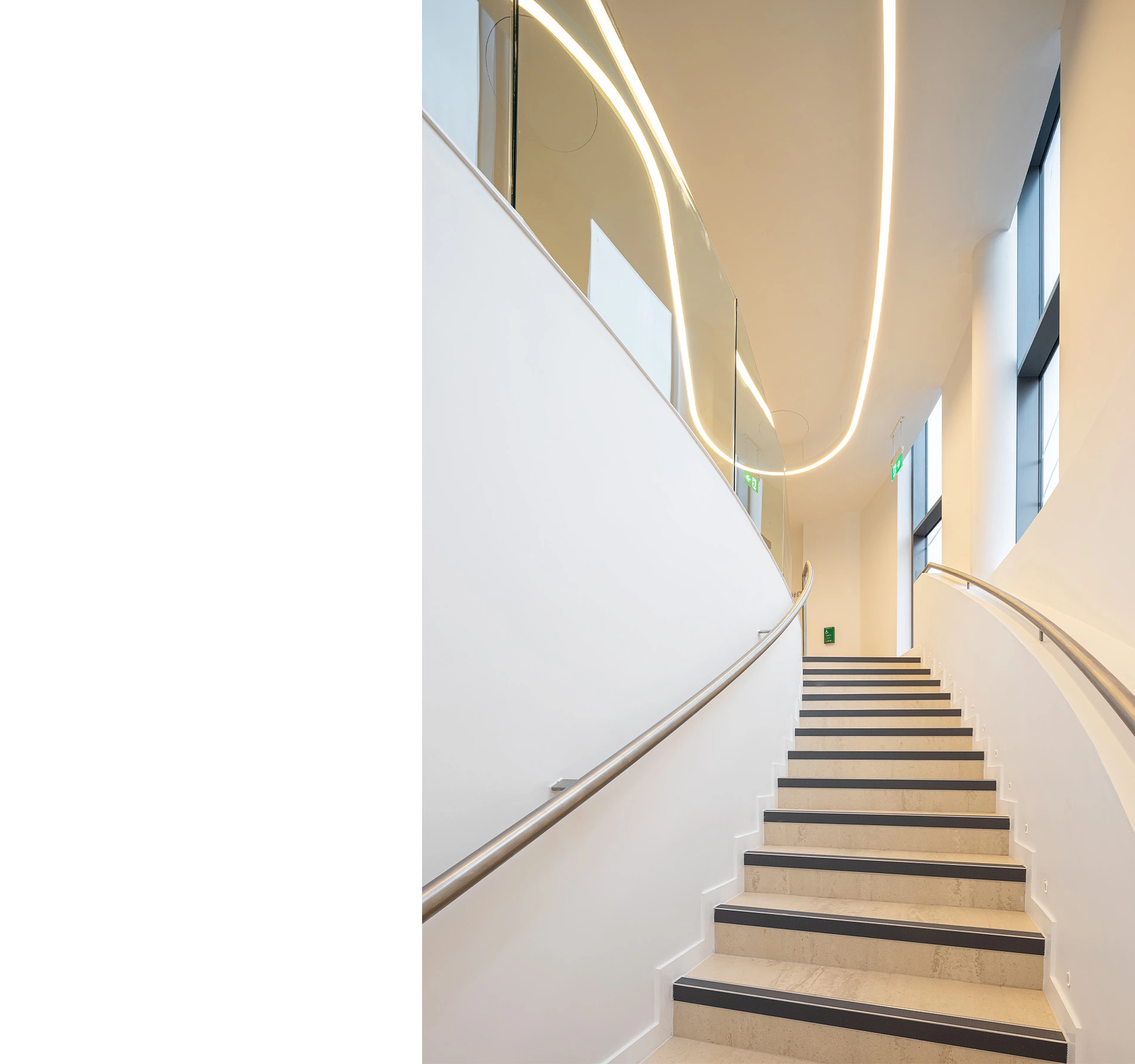 A photo of the main curved staircase leading to the waiting and reception area of a newly opened bespoke contemporary private hospital. The white walls make the space large and welcoming and curved bespoke lights lead the eye to the glass partitioning separating the stairs from the other areas in the open-plan clinic floor. Large windows are visible on the right along the curved wall. This bespoke modern hospital was designed by NVDC healthcare specialist medical architects who have almost two decades of experience with clinic and healthcare architecture premises design.