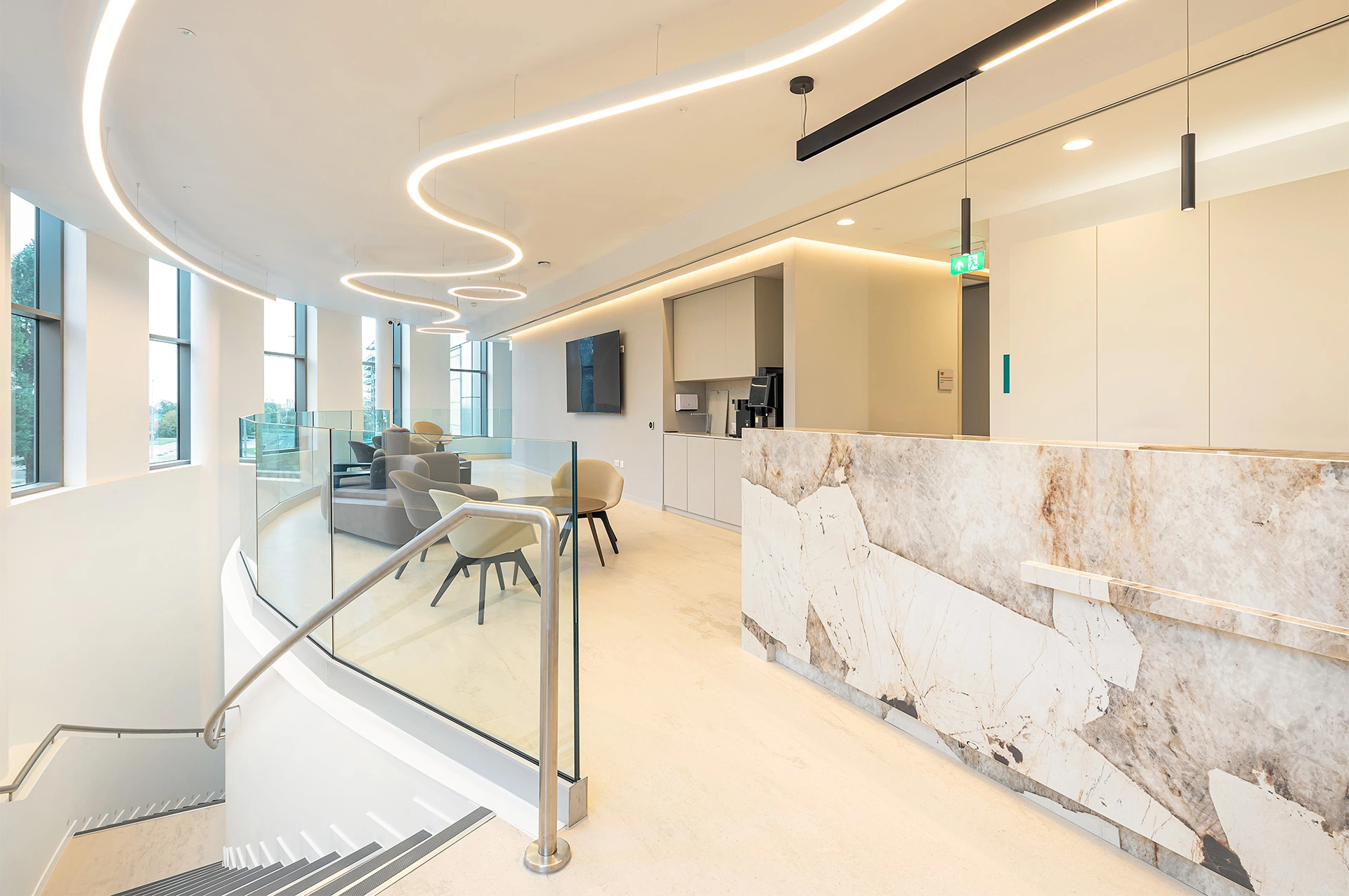 A modern contemporary waiting space and reception on the second floor of a private hospital with the curved stair leading to it visible on the left hand side, large windows and white finished walls. Modern chairs, tables and soft furniture with a small kitchen are provided for the patients to use, and a large TV. Curved bespoke ceiling hanging lights and built-in LEDs along the corners where the walls meet the ceiling drive the onlooker's attention to the corridor on the right hand side leading to the surgery theatres and recovery rooms. A bespoke onyx desk is in the foreground on the right, with light blond stone flooring along the full open-plan space, which is divided by a glass handrail from the rest of the building. This bespoke modern hospital waiting space was designed by NVDC healthcare specialist medical architects who have almost two decades of experience with clinic and healthcare architecture premises design.
