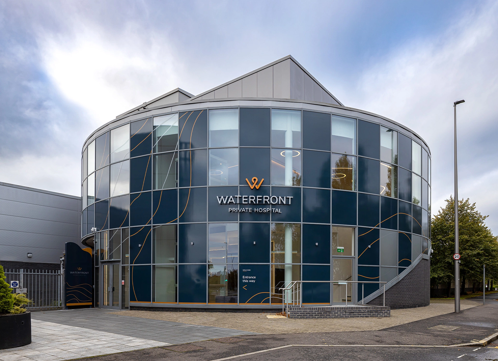 A two-storey building photo of the outside faÃ§ade, which is a curtain wall composed of glazed and coloured dark-blue glass panels, the high floor-to-ceiling windows showcase the hospital waiting room spaces on the inside and the bespoke curved lighting chandeliers. The rest of the building is finished in grey aluminium panels and strips, with an accessibility ramp visible in front of the emergency exit. This bespoke hospital healthcare design was delivered by NVDC Architects, medical specialists in Edinburgh Granton.
