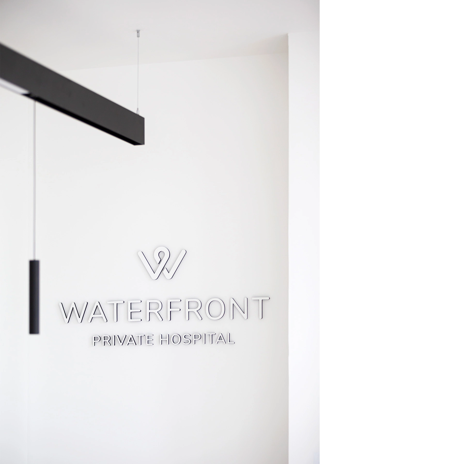 A photo of a white wall with the signage reading Waterfront Private Hospital and a stylish dark grey lighting feature hanging in front of the signage. This bespoke hospital clinic design was delivered by NVDC medical and healthcare specialist architects in Edinburgh Granton, Scotland UK.