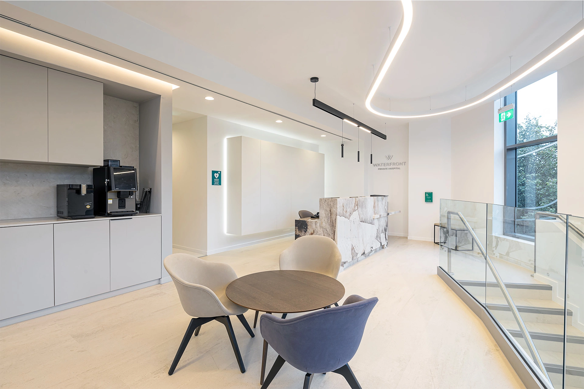 An open-plan space waiting room with a bespoke stone onyx reception desk, a white grey cupboard kitchen with a large coffee machine, and soft chairs with a round table in the foreground. On the right a glass handrail spiral staircase leads to the rest of the hospital. There are curved stylish lighting features hanging from the ceiling, and LEDs behind the small kitchen provided, creating visual highlights throughout the space. Large floor-to-ceiling windows allow for views to the outside and let in lots of natural light. This space was designed to bring comfort while patients wait, and was created and delivered by NVDC medical & healthcare specialists.