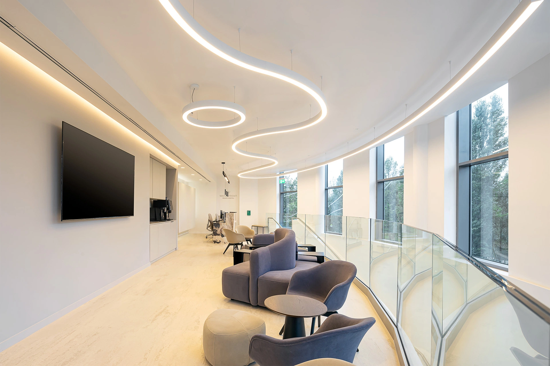 An open-plan space waiting room with a bespoke stone onyx reception desk, a white grey cupboard kitchen with a large coffee machine, and soft chairs with a round table in the foreground. On the right a glass handrail spiral staircase leads to the rest of the hospital. There are curved stylish lighting features hanging from the ceiling, and LEDs behind the small kitchen provided, creating visual highlights throughout the space. Large floor-to-ceiling windows allow for views to the outside and let in lots of natural light. This space was designed to bring comfort while patients wait, and was created and delivered by NVDC medical & healthcare specialists.