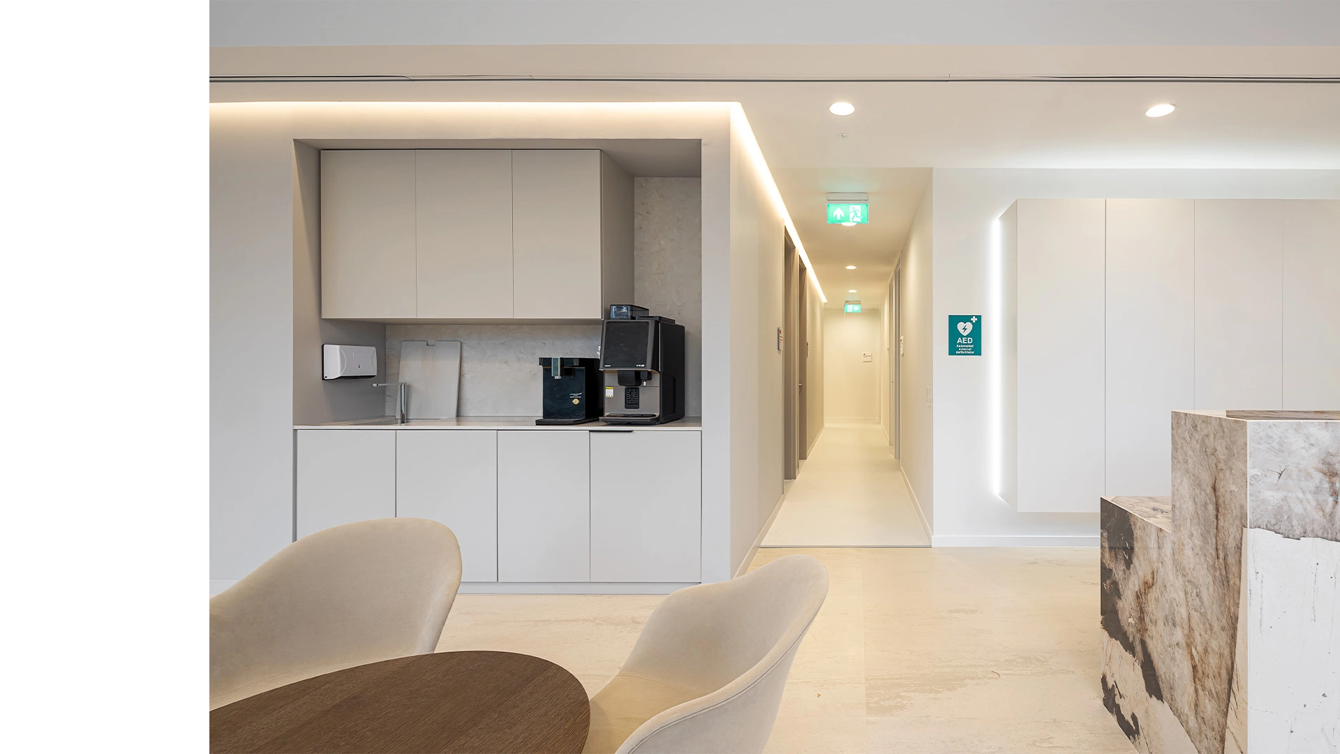 An open-plan space waiting room with a white grey cupboard kitchen with a large coffee machine, and soft chairs with a round table in the foreground. On the right a glass handrail spiral staircase leads to the rest of the hospital. There are curved stylish lighting features hanging from the ceiling, and LEDs behind the small kitchen provided, creating visual highlights throughout the space. This space was designed to bring comfort while patients wait, and was created and delivered by NVDC medical & healthcare specialists.