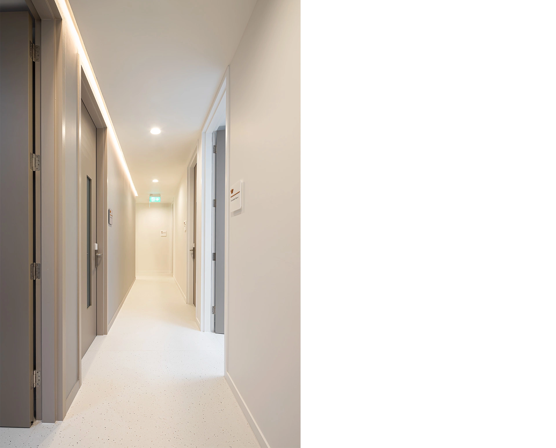 A bright corridor space leading to the surgery theatres and recovery rooms of a recently completed hospital in Edinburgh Granton. The light grey doors, white walls and light stone floors create a warm and inviting atmosphere for patients by reflecting the warms of the bespoke long curving lights and the LEDs placed at the top corners of the rooms. This bespoke hospital private medical premises design was delivered by NVDC Architects who specialise in compliant, safe and patient-centred building design.