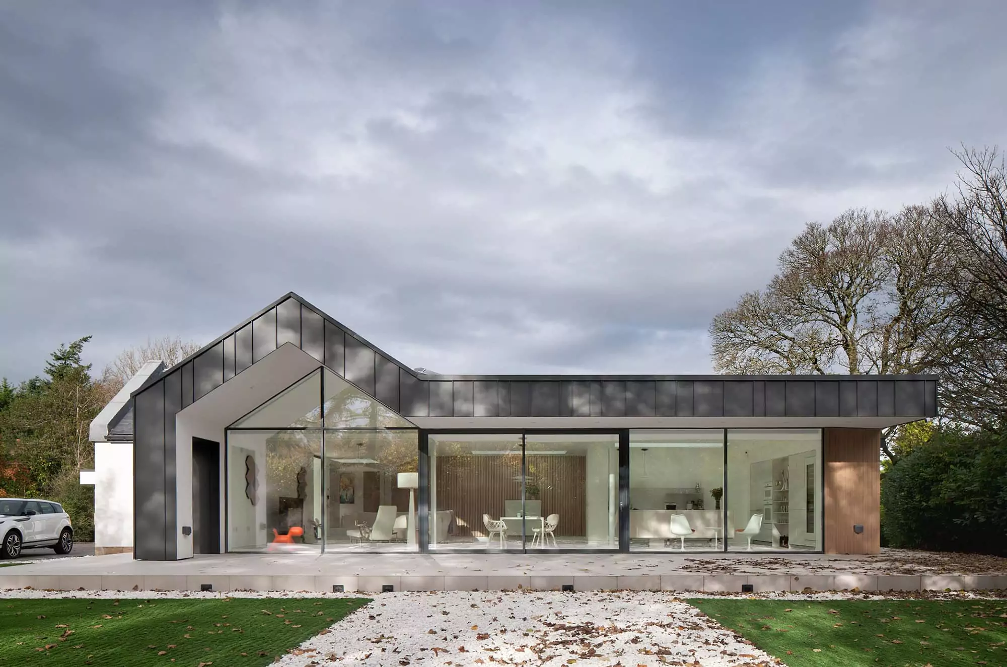 A photo of the exterior of a recently completed retrofit renovation home project in Glasgow, with a protruding veranda roof cladded in black charcoal zinc, and floor-to-ceiling windows showcasing the interior open-plan-living dining and kitchen layout, finished in white and brown timber panel studs. The house has a large garage and a spacious garden from which the shot is taken looking towards the house.
