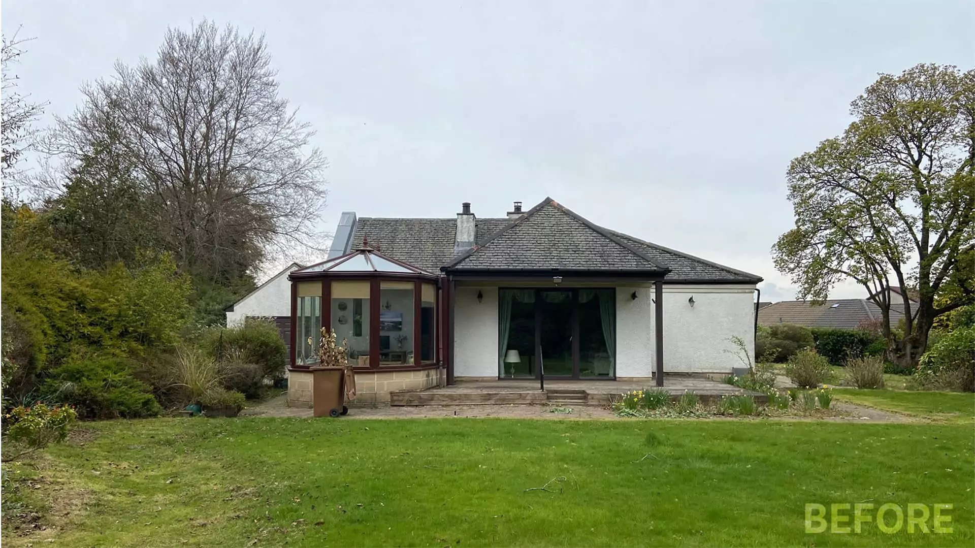 A before photo of a single-storey bungalow house in Glasgow Scotland UK, with slated roof, white render exterior walls and a round bay window on the left side. The garden surrounding the house has nice luscious bushes and a bright green lawn.
