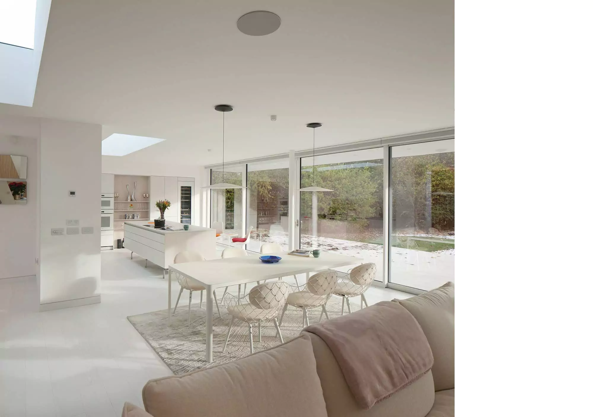 A photo showing the interior design and décor of a recently completed bespoke house project. The walls and ceiling are finished in white plaster, with white timber floors, and large rooflights glowing in the hazed cloudy UK weather. Both the kitchen and the open-plan-living and dining are finished in white, with white soft furniture, chairs, tables, kitchen island, and wine cooler. The floor-to-ceiling windows allow lots of natural light in and serve to showcase views towards the large garden. This project was designed by NVDC Architects specialists in house improvement and remodelling Scotland and the UK.