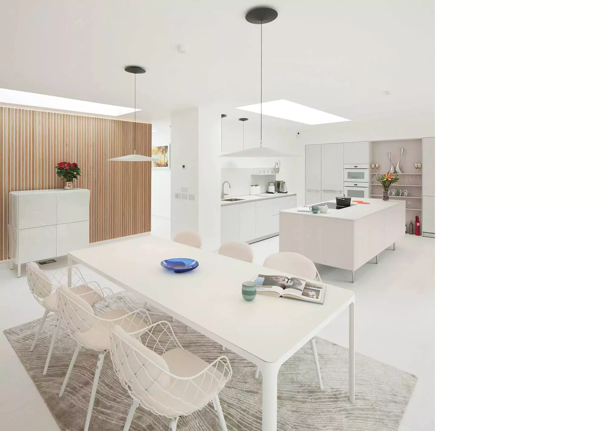A photo of the interior of an all-white kitchen living and dining space with a white kitchen island, and all white kitchen cabinets and countertops, including a white pantry wall and a white table and chairs in the forefront. There are two rooflights visible which allow for natural light into the deep space of the house extension - one above the kitchen space and one next to the back timber panel finished wall. This project was designed and delivered by NVDC Architects residential design experts.
