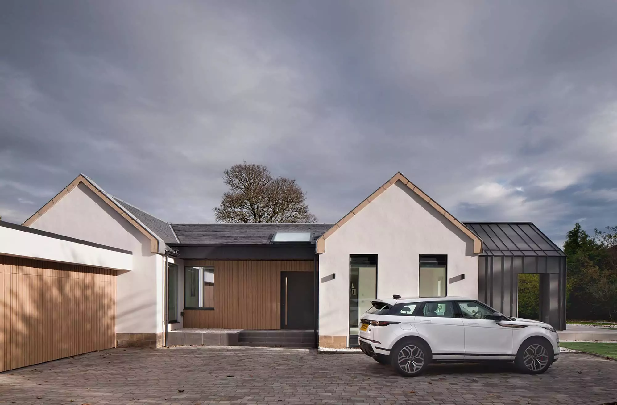 A photo of the side of a single-storey house extension in Glasgow Newton Mearns Scotland UK. There are two gables facing the photo and one extruding where the zinc panel cladded overhang creates shelter for the extended house veranda. The entrance and garage are finished in timber panels which create visual interest of textures, some blond sandstone details of the original house were kept on the corners and where the house meets the ground. The roof is finished in grey slates and there is a rooflight window visible right above the entrance door. This house extension and refurbishment retrofit project was delivered by NVDC Architects, experts in home renovation and remodelling.