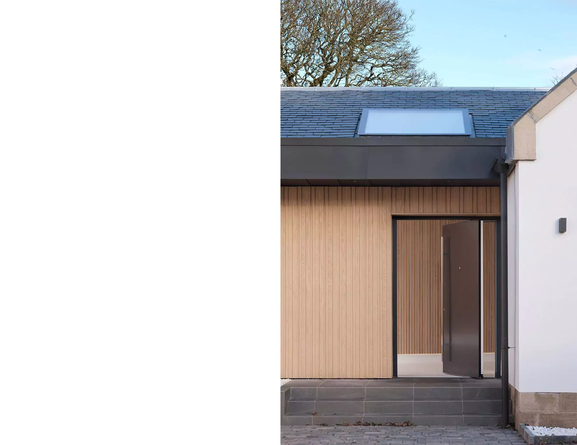 Modern luxurious house entrance with a large aluminium door in charcoal black and timber batten finished acoustic panel walls on the exterior and interior visible through the open door. The flashings are dark zinc, which goes well tonally with the dark grey slated roof. There is a rooflight visible right above the entrance.