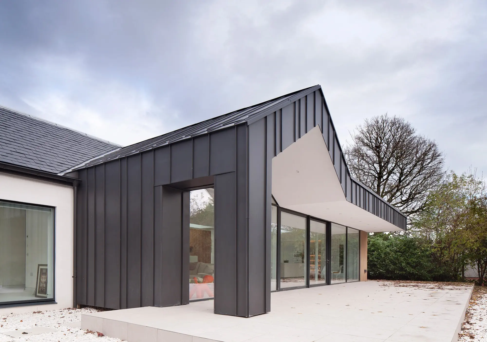 Contemporary house extension in Glasgow featuring sleek zinc cladding and expansive floor-to-ceiling windows, designed by Glasgow architects specializing in modern remodelling and renovations. This architectural project emphasises open-plan living, maximising natural light through large windows and creating a seamless blend between indoor and outdoor spaces, showcasing the firm's expertise in innovative exterior design solutions. the zinc cladded overhang shelters the white stone tile finished veranda creating the opportunity for the clients to enjoy their garden while protected from the weather.