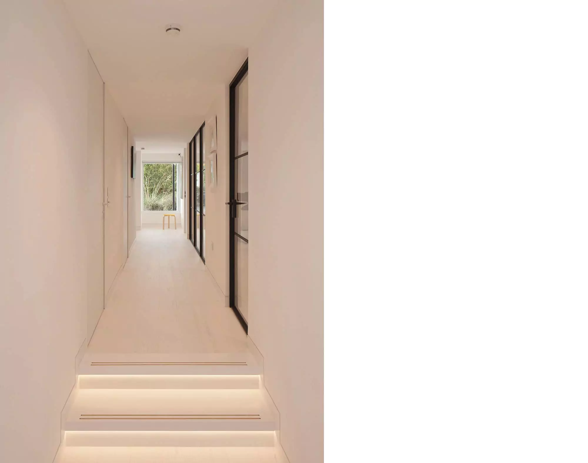 Elegant hallway area designed by Glasgow architects, emphasizing the harmony of light and space in this house extension. The use of white interiors, zinc cladding, and strategic rooflights brings in an abundance of natural light, while LED lighting under the steps leading to the corridor subtly enhances the ambiance. The carefully crafted renovation and remodelling work exemplify modern interior design, ideal for clients seeking to transform their homes into modern luxurious spaces which respond to their contemporary lifestyle. The hallway ends with a large window looking towards the luscious green bushes in the house garden outside with a small chair and a seating space on the window sill providing a minimalist reading nook.