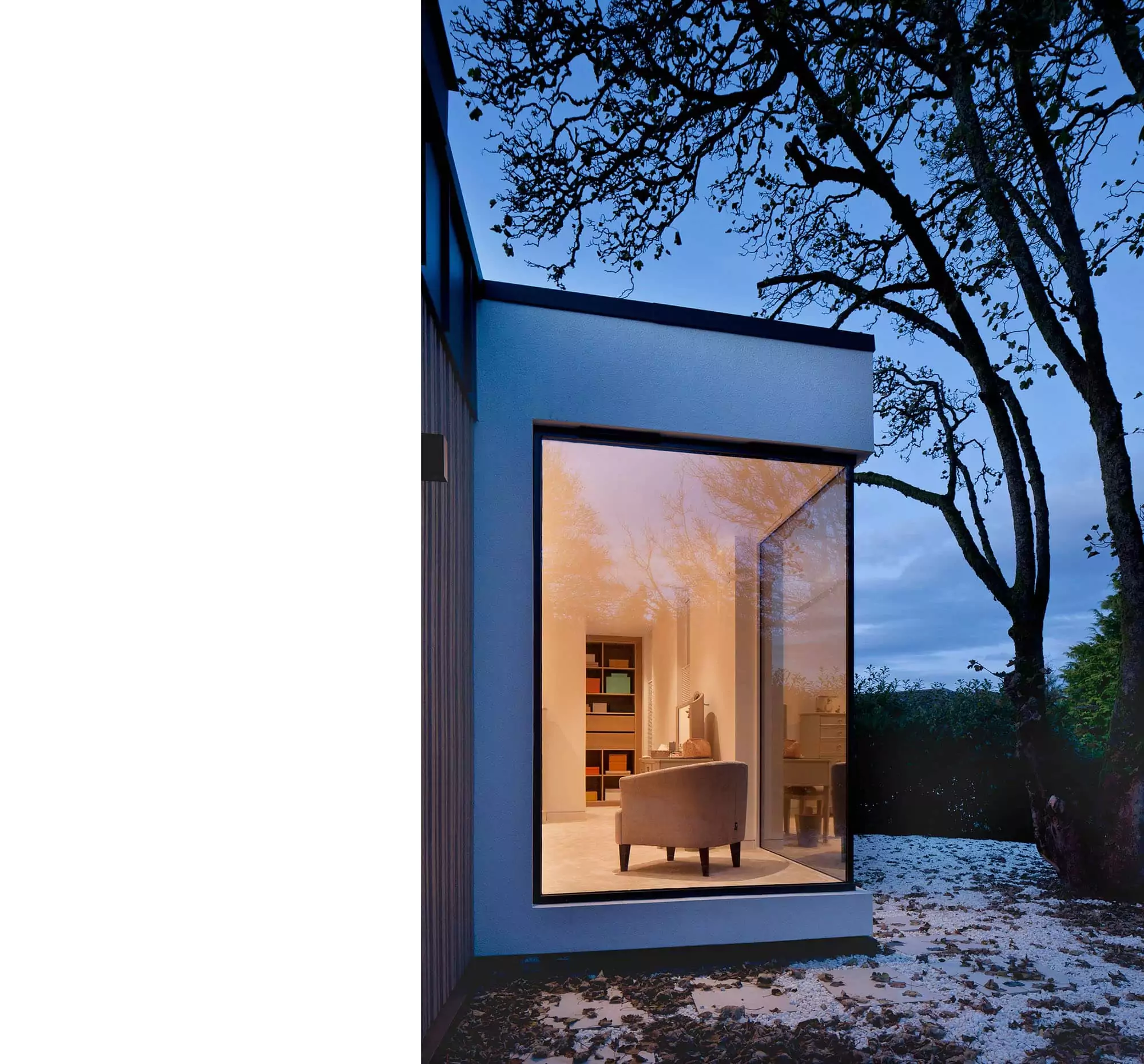 Dusk exterior of a house extension designed by Glasgow architects, featuring a large floor-to-ceiling angle window that offers a view into the bespoke bedroom space, bathed in warm light. The remodelling project incorporates a rooflight and zinc cladding, emphasizing a modern aesthetic. The well-lit interior reveals a tastefully furnished space with white walls, creating a bright and airy atmosphere. This renovation showcases the seamless integration of indoor and outdoor spaces, emphasizing natural light and contemporary architectural design, specifically designed to allow clients to enjoy their garden and outdoor premises more from the comfort of their own bedroom.